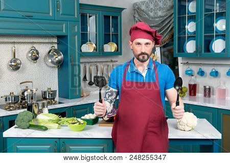Brave Chef With Spoons In Hands In Red Apron And Red Cap. Guy In Hat And Apron Standing On Kitchen I