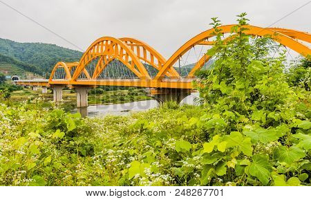 Landscape Of Yellow Arched Railroad Bridge Spanning A River In The Countryside On A Cloudy Overcast 