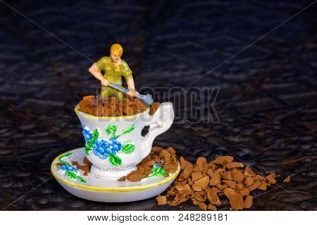A Tiny Miniature Male Worker Figure Shoveling Coffee Grains Out Of A Miniature Tea Cup And Saucer
