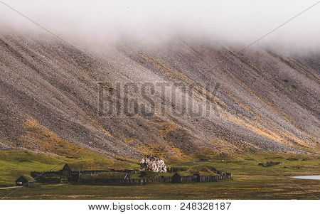 Old Viking Village In Iceland With Foggy Hill. Old Wooden Buildings Covered Grass