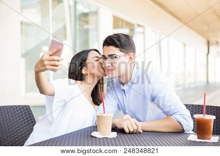 Teenage Girl Kissing Boyfriend On Cheek While Taking Selfportrait On Mobile Phone In Shopping Mall