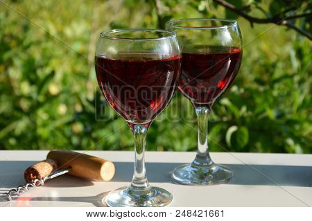 Red Wine Poured Into Wine Glass On Background Of Green Tree Leaf Foliage. Purpie Flower