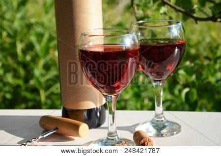 Red Wine Poured Into Wine Glass On Background Of Green Tree Leaf Foliage. Purpie Flower