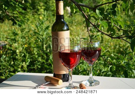 Red Wine Poured Into Wine Glass On Background Of Green Tree Leaf Foliage. Purpie Flower