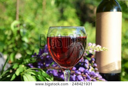 Red Wine Poured Into Wine Glass On Background Of Green Tree Leaf Foliage. Purpie Flower