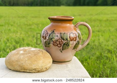 Still Life With A Ceramic Jug And Loaf Of Rustic Bread On A Tablecloth Against The Background Of A G