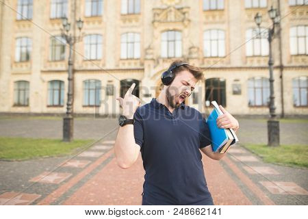 Happy Man With Books In His Hands Listening To Music In Headphones Against The Background Of The Uni