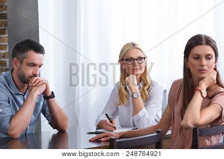 Woman Turned Away And Her Frustrated Husband Sitting At Table On Therapy Session By Female Counselor