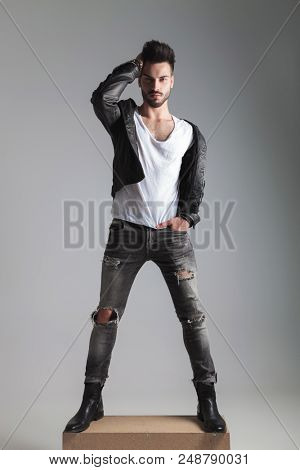 handsome man in leather jacket fixing his hair while standing on a wooden box with a hand in pocket, full length picture