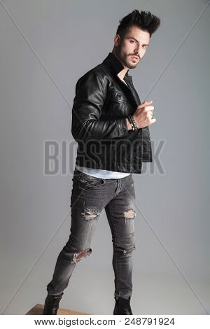 handsome man holding his leather jacket turns to side while standing on a wooden crate on light grey background, full body picture