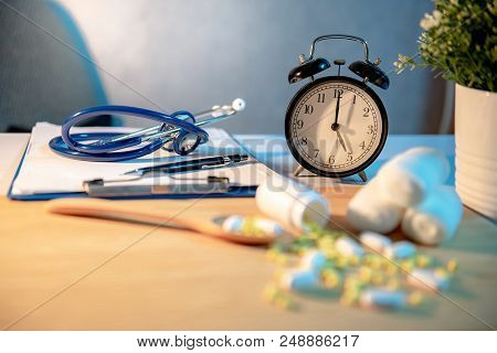Stethoscope, Clipboard, Pen And Table Clock On Dector Desk. Blurred Pills Spilling Out Of Pill Bottl