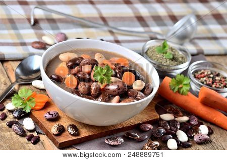 Bowl Of Bean Soup With Large Beans On Cutting Board, Carrots, Parsley, Marjoram, Spoon, Towel And La