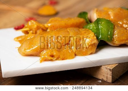 closeup of some raw spiced chicken meat skewers, mixed with green pepper, on a white marble tray, on a wooden table