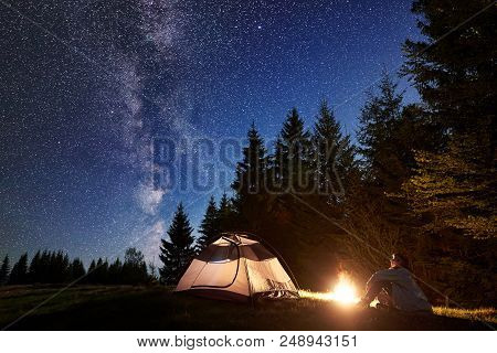 Young Man Sitting Alone In Front Of Tourist Tent At Burning Bonfire On Grassy Valley Enjoying Dark B