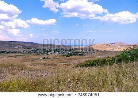 A Landscape Of Vague Gray Hills And A Settlement In A Valley With A Narrow Strip Of Sea Blue In The 