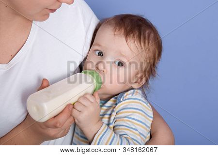 Hirizontal Shot Of Liitle Charming Baby Drinking Milk Of Bottle, Being In Mother's Arms, Feels Hunge