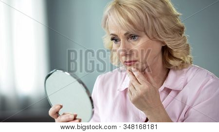Unhappy Senior Female Looking At Sagging Skin Face In Mirror, Old Age Appearance