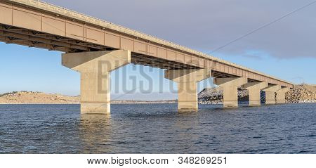 Stringer Bridge Spanning Over A Lake With View Of Snowy Terrain And Cloudy Sky