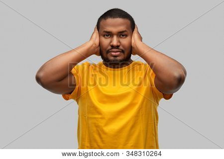 stress, noise and people concept - stressed young african american man in yellow t-shirt closing his ears with hands over grey background