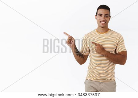 Good-looking Modern Young Man With Mascules, Tattoo, Pointing Upper Left Corner And Smiling Happily,