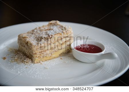 A Piece Of Napoleon Cake On A White Plate. The Cake Is Sprinkled With Powdered Sugar. There Is A Cup