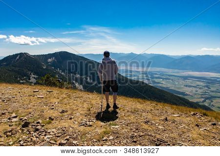 A Young Man Holding A Crutch And Hiking. A Beautiful Hilltop Landscape. Extremal Hiker Looking At Th