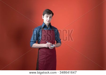 Handsome Barista In Blue Shirt With Rolled Sleeves And Red Apron Writing In Notebook And Looking At 