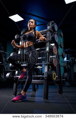 From Below View Of Female Bodybuilder Posing Near Stand With Sports Equipment, Touching Braids. Srto