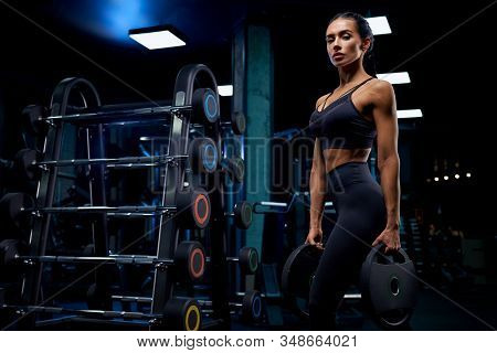 Side View Of Fitnesswoman With Braids Posing Near Stand With Sports Equipment And Holding Weights. S