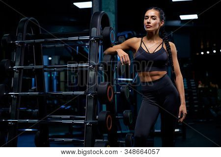 Front View Of Female Bodybuilder With Long Braids Posing Near Stand With Sports Equipment. Srtong Wo