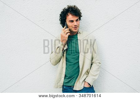 Smart Young Man Smiling And Talking On Mobile Phone With His Friend Against White Cocnrete Wall. Han