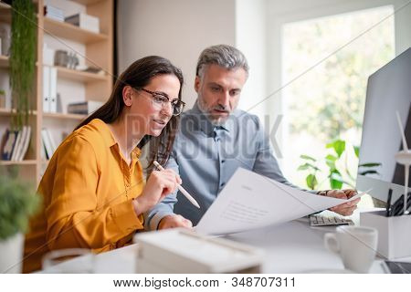 Architects Sitting At The Desk Indoors In Office, Looking At Blueprints.