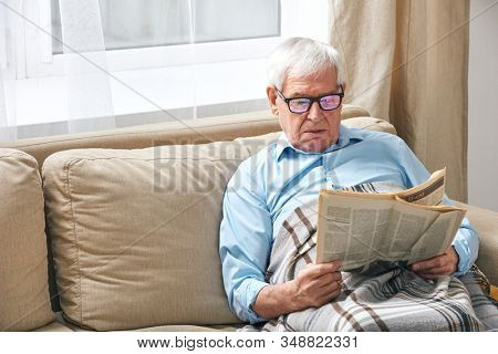 Senior grey-haired man wrapped in plaid reading newspaper while relaxing on couch by window while staying at home
