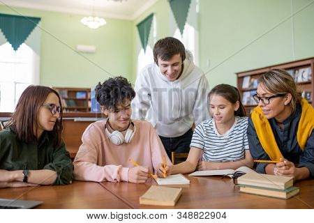 Large group of teenage learners sitting by desk in college library while discussing points of forthcoming seminar