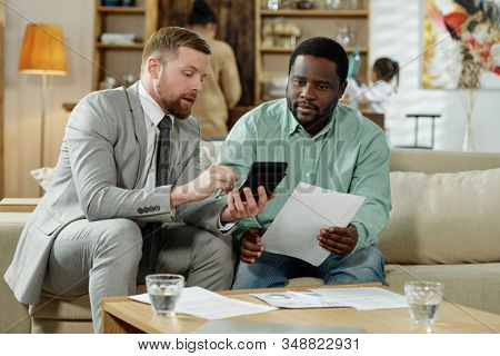 Adult black man with professional adviser sitting on couch with calculating machine discussing real estate mortgage