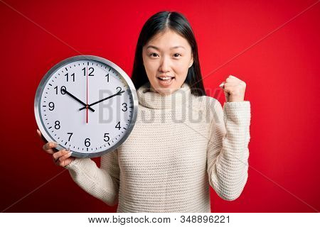 Young asian woman holding countdown big clock over red isolated background screaming proud and celebrating victory and success very excited, cheering emotion