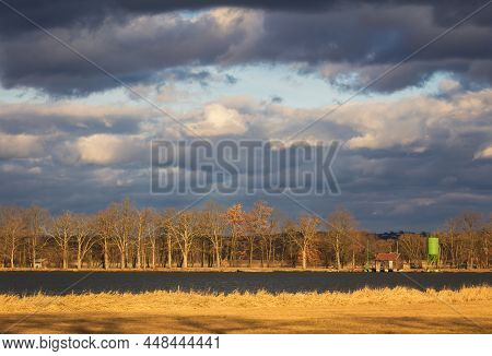 Dramatic Sunset Sky Above Pond With Fishing Building, Granary And Leafless Tree Aley On Water Edge. 