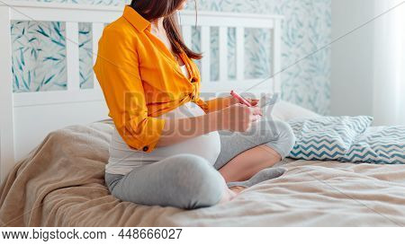 Pregnant Woman Writing Down In Notebook In Bed On Bright Background.
