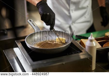 Crope View Of Chefs Hands In Black Gloves Melting Piece Of Butter In Frying Pan. Male In Chef Unifor
