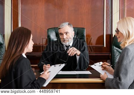 Senior Judge Holding Eyeglasses Near Attorney And Prosecutor Writing On Clipboards