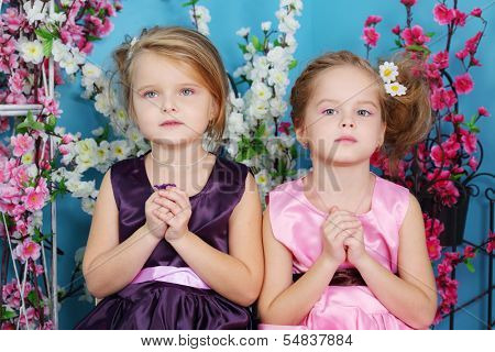 Two cute little girls in dresses sit with folded hands in prayer in room with flowers.