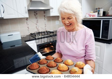 A laughing grandmother holding a tray of baked muffins just out of the oven
