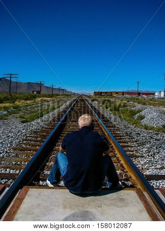 A lonely blond boy in a dark blue blouse, is sitting back to the camera on the grade crossing, looking ahead towards the railway track and surrounding grasslands.