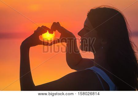 Beautiful female model enjoying sunset and making heart sign on sun at seaside. Calm water of salt lake Elton reflects silhouette of woman. Sun goes under horizon. Girl stands alone in water. Sun reflects in Elton's water.