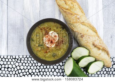 Courgette soup with sour cream in black bowl . Sliced zucchini and long fresh baguette on the white wooden background. Top view