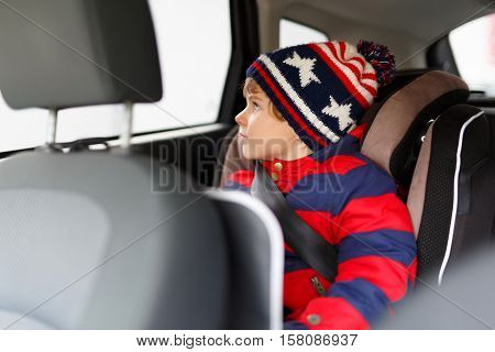 Adorable cute preschool kid boy sitting in car in winter clothes. Little child in safety car seat with belt enjoying trip and jorney. Safe travel with kids and traffic laws concept.