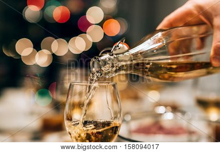Closeup of a man pouring white wine into a glass on a dining table full of food with a Christmas tree in the background