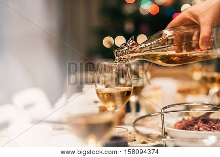 Closeup of a man pouring white wine into a glass on a dining table full of food with a Christmas tree in the background