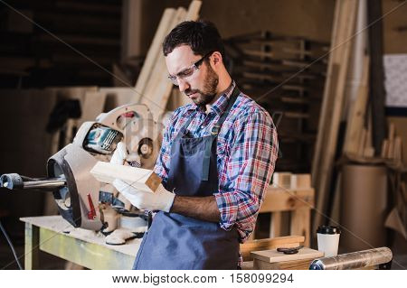 Handsome carpenter in protective glasses standing near his wooden object in the workshop.