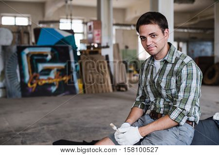 Handsome carpenter in protective glasses is looking at camera and smiling while standing near his wooden object in the workshop.